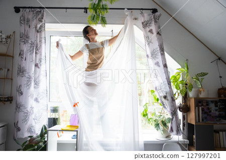 Woman in apron hangs transparent tulle curtains on large attic windows in the house inside the interior with potted plants. Spring cleaning, tidying up Woman in apron hangs transparent tulle curtains on large attic windows in the house inside the interior with potted plants. Spring cleaning, tidying up 127997201