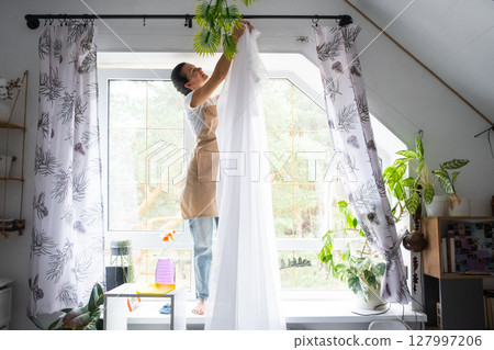 Woman in apron hangs transparent tulle curtains on large attic windows in the house inside the interior with potted plants. Spring cleaning, tidying up Woman in apron hangs transparent tulle curtains on large attic windows in the house inside the interior with potted plants. Spring cleaning, tidying up 127997206