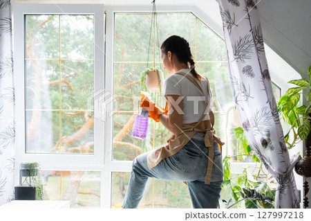 Woman in rubber gloves and apron manually washes attic window of house with rag cleaner and mop inside interior with home plants. Restoring order and cleanliness in spring, cleaning servise 127997218