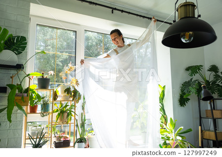 Woman in apron hangs transparent tulle curtains on large windows in the house inside the interior with potted plants. Spring cleaning, tidying up Woman in apron hangs transparent tulle curtains on large windows in the house inside the interior with potted plants. Spring cleaning, tidying up 127997249