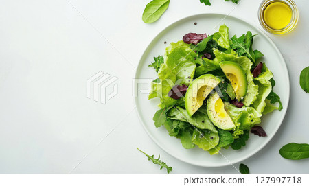 Fresh green salad with sliced avocado, mixed leafy greens, black pepper and olive oil on white plate over light background. Healthy clean eating. 127997718