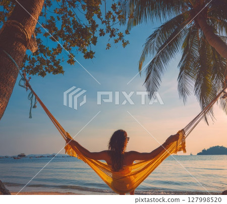 Tropical Sunset Serenity: Woman Relaxes in a Hammock on a Secluded Beach, Embracing Peaceful Island Life and Golden Hour Views 127998520
