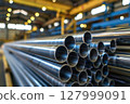 Stack of polished metal pipes inside a modern industrial warehouse, focus on steel texture. 127999091