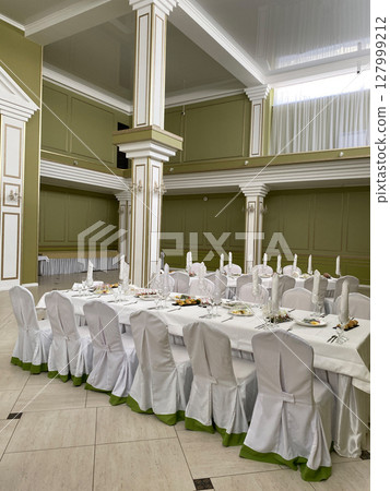 Banquet table setup in an elegant hall with white covered chairs and tall columns. Prepared plates, folded napkins, and crystal glassware create a refined celebration atmosphere 127999212