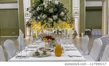 Elegant banquet table with floral centerpiece and fruits, set for a formal celebration. Glassware, cutlery, and a pitcher of juice complement the luxurious event setup in beautiful venue Elegant banquet table with floral centerpiece and fruits, set for a formal celebration. Glassware, cutlery, and a pitcher of juice complement the luxurious event setup in beautiful venue 127999217