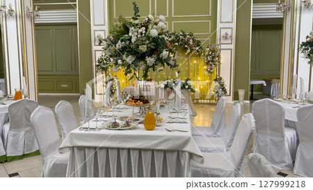 Elegant banquet table with floral centerpiece, fresh fruit, and drinks in luxurious banquet hall. White chair covers, glasses, and fine dining set the scene for a formal celebration or wedding 127999218
