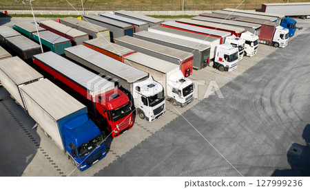 Aerial view of a large truck parking lot at a logistics hub, with multiple commercial semi trucks parked in organized rows. Aerial view of a large truck parking lot at a logistics hub, with multiple commercial semi trucks parked in organized rows. 127999236