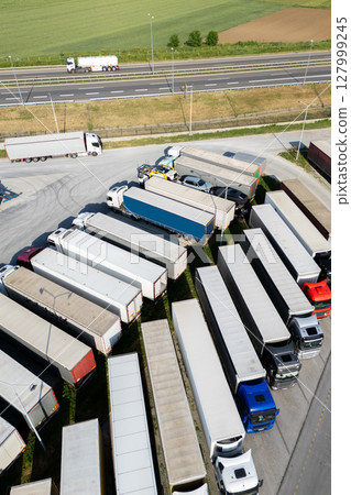 Aerial view of a large truck parking lot at a logistics hub, with multiple commercial semi trucks parked in organized rows. Aerial view of a large truck parking lot at a logistics hub, with multiple commercial semi trucks parked in organized rows. 127999245