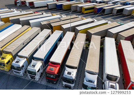 Aerial view of a large truck parking lot at a logistics hub, with multiple commercial semi trucks parked in organized rows. Aerial view of a large truck parking lot at a logistics hub, with multiple commercial semi trucks parked in organized rows. 127999253