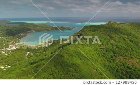 Tropical vegetation covers the mountain slopes of Bora Bora, a volcanic island in French Polynesia, while turquoise waters lap against the sandy shores of the lagoon 127999456