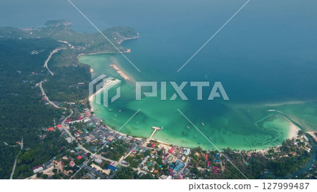 Aerial view capturing a coastal road winding through lush greenery, leading to a vibrant town beside a picturesque bay with crystal-clear turquoise waters on Koh Kood island 127999487