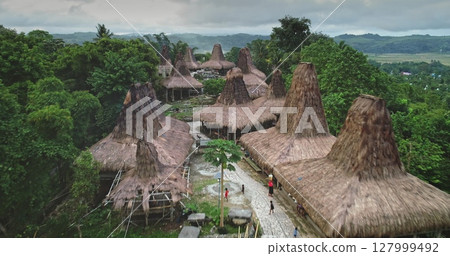 Aerial view of unique custom houses with high, thatched roofs forming a captivating ancient village in a remote area of Sumba island, Indonesia, with local people walking on a path Aerial view of unique custom houses with high, thatched roofs forming a captivating ancient village in a remote area of Sumba island, Indonesia, with local people walking on a path 127999492