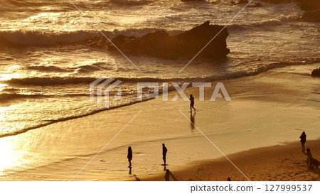 Lisbon, Portugal: Golden sunlight reflecting on wet orange sand, tourists people walk along the sunset beach, creating a warm and inviting relaxing joy atmosphere. Summer travel vacation background 127999537