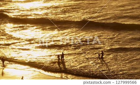 Lisbon, Portugal: Golden waves washing sandy beach, orange sun light reflecting on water, tourists people enjoying sunset over Atlantic Ocean, creating idyllic scene. Summer travel vacation background Lisbon, Portugal: Golden waves washing sandy beach, orange sun light reflecting on water, tourists people enjoying sunset over Atlantic Ocean, creating idyllic scene. Summer travel vacation background 127999566