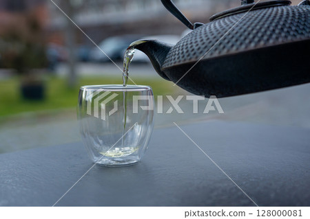 Pouring green tea into a glass from a metal kettle on blurred background, hot drink in teapot 128000081