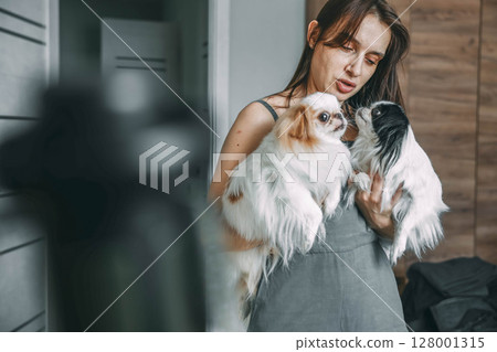 woman tenderly holding two japanese chin dogs, showcasing affection and pet care at home 128001315