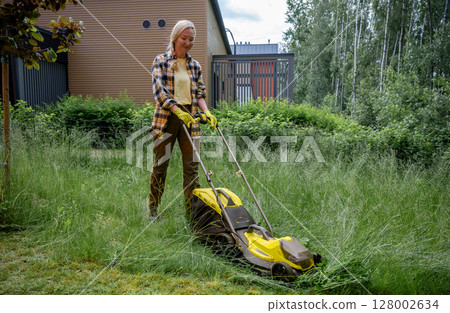 Woman using electric lawn mower in overgrown garden near wooden house Woman using electric lawn mower in overgrown garden near wooden house 128002634