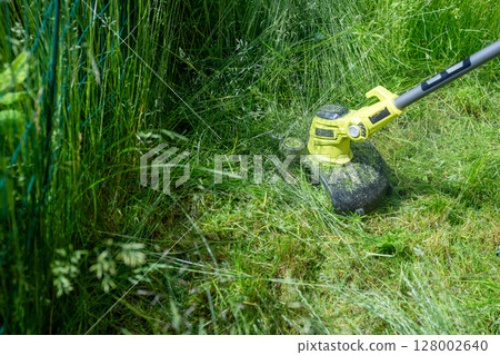 man trimming overgrown grass with electric string trimmer in summer garden 128002640