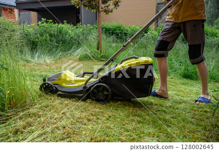 Man mowing overgrown lawn with yellow electric mower near wooden house steps 128002645