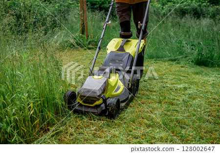 Man mowing overgrown lawn with yellow electric mower near wooden house steps 128002647