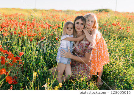 Happy mother with two daughters enjoying picnic in blooming poppy field. Family love concept. 128005015