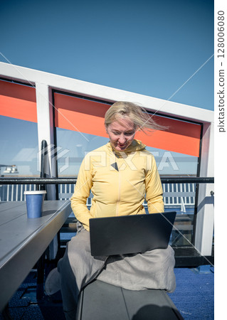 Woman working on laptop outdoors on ferry deck with coffee on sunny windy day 128006080