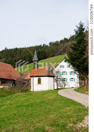 Charming village landscape featuring a quaint chapel and traditional houses nestled among rolling green hills on a serene spring day 128006784