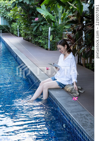 Asian woman reading book relaxing by the poolside, dipping feet in the turquoise water, use hand stroking cat for play with domestic animal on vacation day. Animal therapy, Life with Pets concept Asian woman reading book relaxing by the poolside, dipping feet in the turquoise water, use hand stroking cat for play with domestic animal on vacation day. Animal therapy, Life with Pets concept 128007861