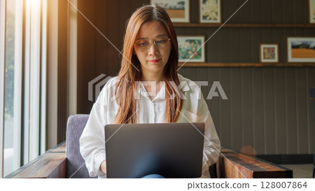 working from home, remote work concept. front view of Asian woman in casual shirt wearing eyeglasses using laptop computer for work at home. morning light flare from window working from home, remote work concept. front view of Asian woman in casual shirt wearing eyeglasses using laptop computer for work at home. morning light flare from window 128007864