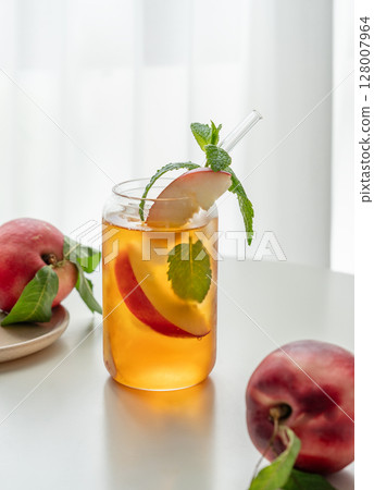 Ice tea with peach, mint in a glass on a white table near window with fresh fruits and sunlight. 128007964