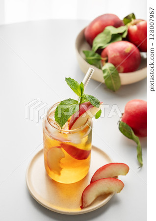 Ice tea with peach, mint in a glass on a white table near window with fresh fruits and sunlight. 128007967