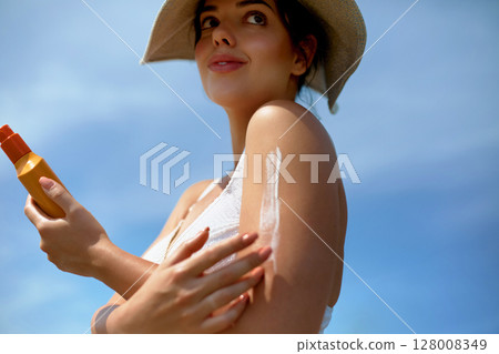 A stylish woman wearing a hat applies sunscreen lotion to her shoulder on a sunny day. The concept conveys skin care, sun protection, and enjoying outdoors in bright, warm weather A stylish woman wearing a hat applies sunscreen lotion to her shoulder on a sunny day. The concept conveys skin care, sun protection, and enjoying outdoors in bright, warm weather 128008349