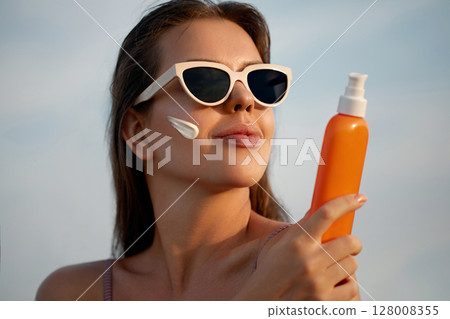 A beauty woman applying sunscreen on healthy skin. Portrait of a female holding a bottle of sun cream and using cream. Skincare product, suntan lotion, or moisturizing solar A beauty woman applying sunscreen on healthy skin. Portrait of a female holding a bottle of sun cream and using cream. Skincare product, suntan lotion, or moisturizing solar 128008355