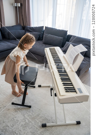 Little girl preparing to play piano at home in the living room. Calm moment of focus and anticipation before music practice in a cozy family space. Little girl preparing to play piano at home in the living room. Calm moment of focus and anticipation before music practice in a cozy family space. 128008724