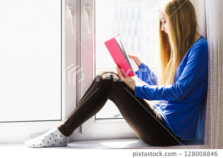 Woman sitting on window sill reading book at home 128008846