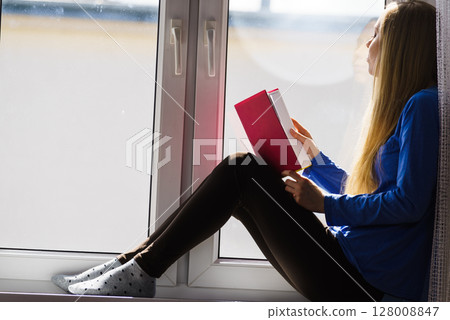Woman sitting on window sill reading book at home Woman sitting on window sill reading book at home 128008847