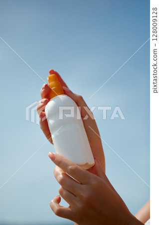 Close-up of a person's hands holding a sunscreen bottle with SPF, with a vivid blue sky in the background, evoking themes of summer, skincare, and outdoor relaxation Close-up of a person's hands holding a sunscreen bottle with SPF, with a vivid blue sky in the background, evoking themes of summer, skincare, and outdoor relaxation 128009128