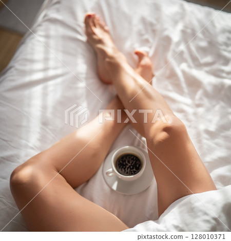 Close-up of female legs with cup of coffee on bed. Close-up of female legs with cup of coffee on bed. 128010371