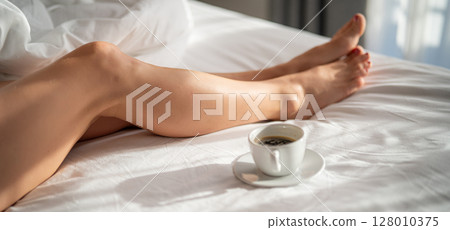 Close-up of female legs with cup of coffee on bed. 128010375