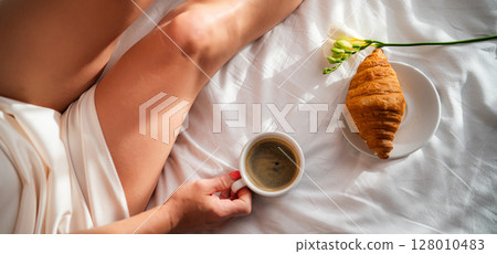 Close-up of female legs with cup of coffee on bed. 128010483