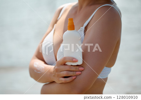 Close-up of a woman in a white bikini holding a sunscreen bottle at a sunny beach 128010910