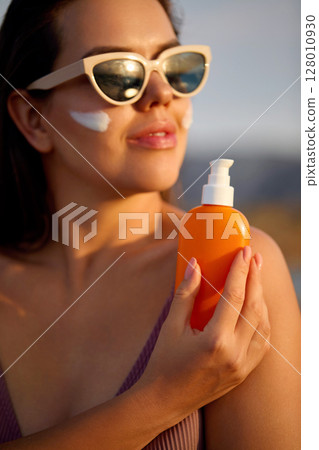 A beauty woman applying sunscreen on healthy skin. Portrait of a female holding a bottle of sun cream and using cream. Skincare product, suntan lotion, or moisturizing solar. A beauty woman applying sunscreen on healthy skin. Portrait of a female holding a bottle of sun cream and using cream. Skincare product, suntan lotion, or moisturizing solar. 128010930