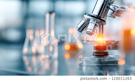 Close-up of a modern microscope with illuminated stage, surrounded by laboratory glassware, showcasing scientific research and innovation in a lab environment 128011263
