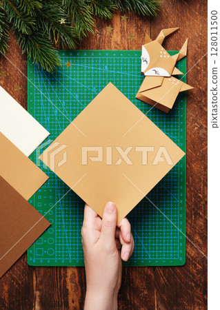 A hand holds a square sheet of brown paper for origami. In the background is a workspace for paper crafts. A cutting mat, origami paper, pencils, and a ruler. 128011500