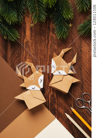 Two deer, handmade from brown paper using origami techniques, on a wooden surface surrounded by evergreen branches, gold star confetti, and art supplies. 128011506