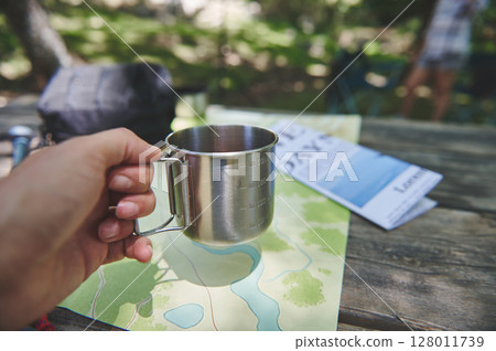 Camping Setup Featuring a Stainless Steel Mug on a Wooden Table with Maps and Guides 128011739