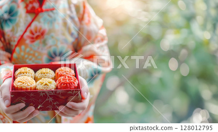 Close up of child in traditional chinese kimono holding festive red box with colorful mooncakes. Banner for mid autumn festival, celebration and cultural traditions. Copy space for text 128012795