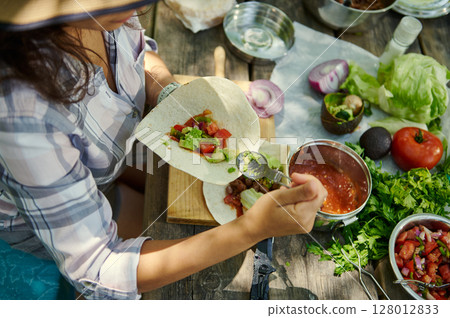 Woman Preparing Fresh Tacos Outdoors with Vibrant Ingredients and Herbs 128012833