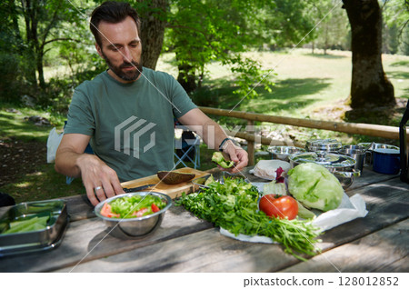 Man Preparing Fresh Salad at Outdoor Picnic Table Surrounded by Nature 128012852