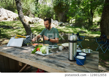 Man Preparing Food at an Outdoor Picnic Table in a Forest Setting 128012858